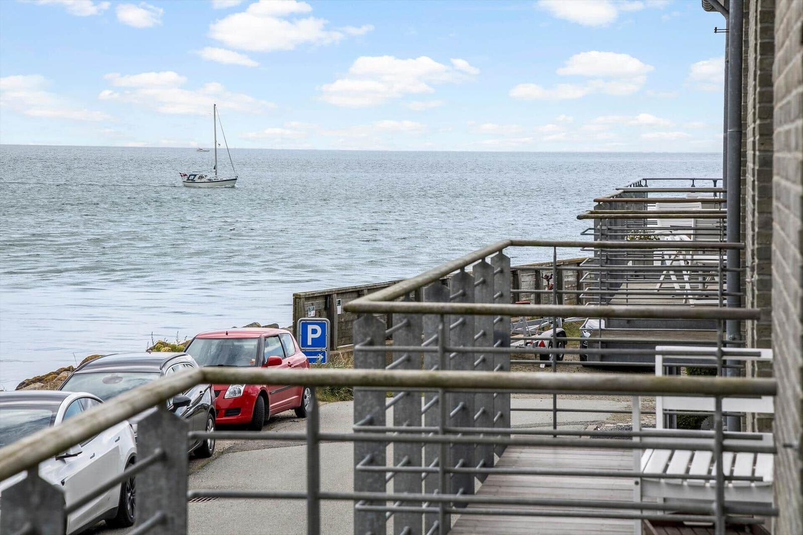 Meerblick mit Segelyacht, Parkplatz und Holzterrasse mit Metallgeländer.