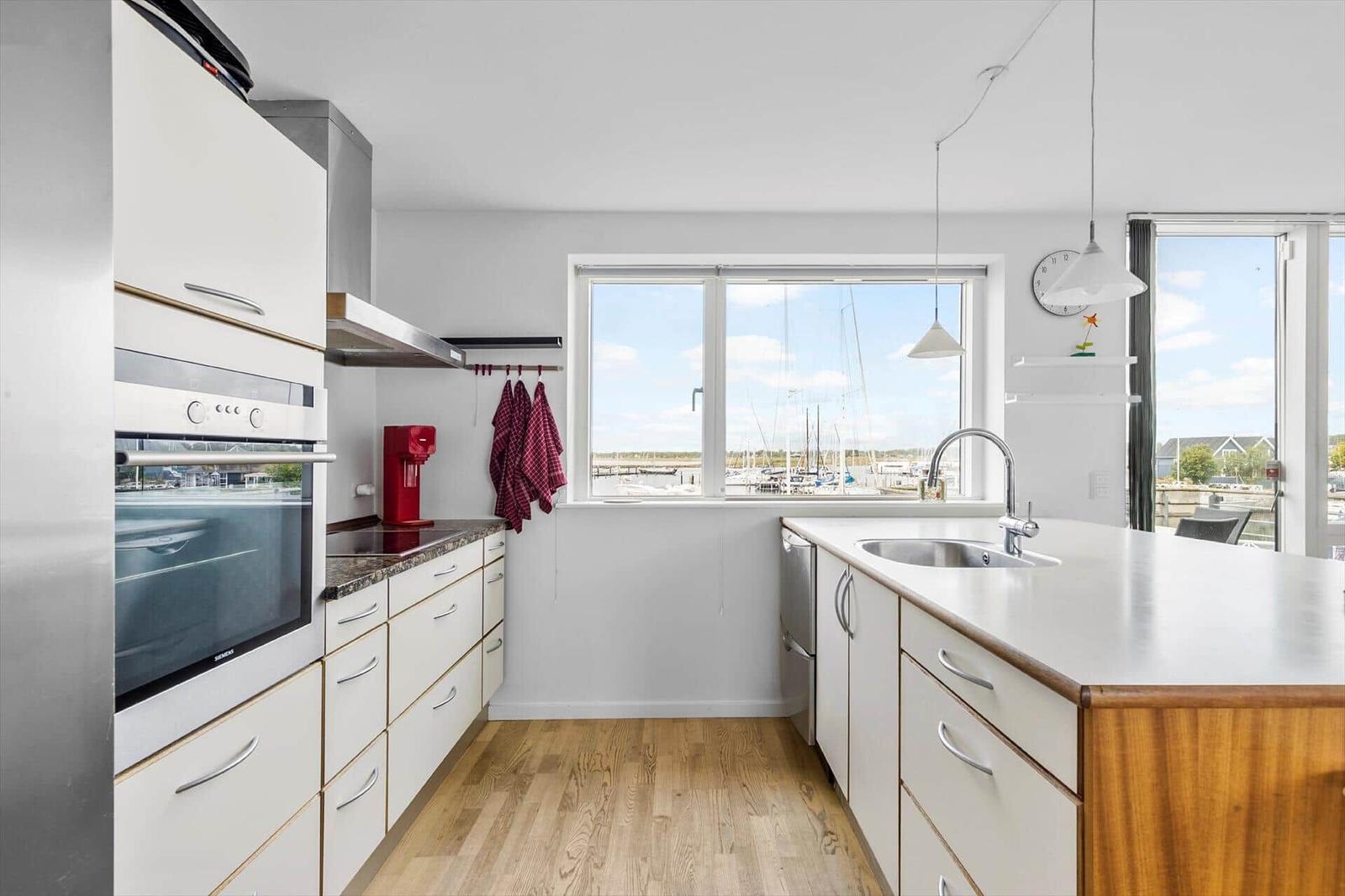 Küche mit Blick auf Hafen und Yachten. Weiße Schränke, Holzboden und Edelstahlgeräte.