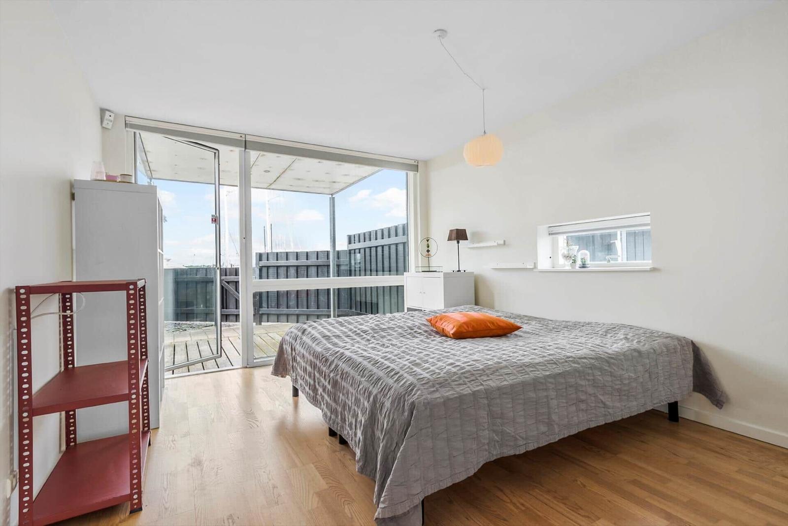 Schlafzimmer mit Balkon, Bett und Holzboden. Fenster und Tür bieten Blick auf Außenbereich.