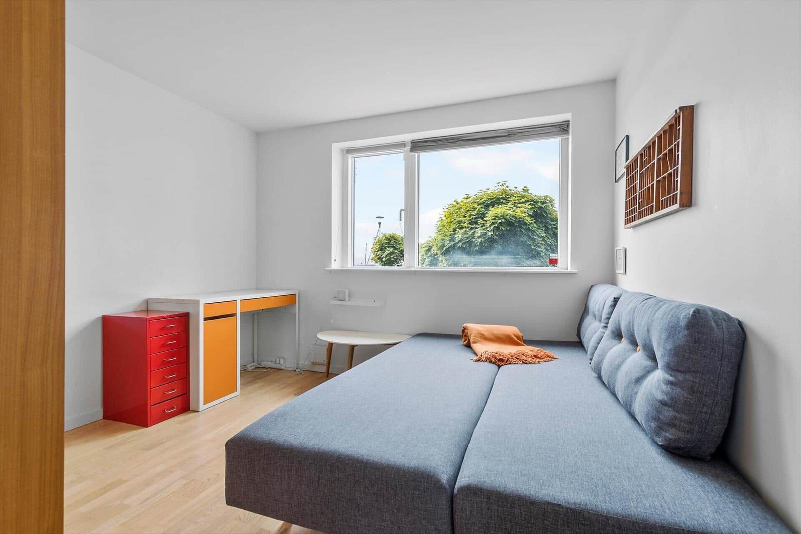 Ein Zimmer mit blauem Sofa, Schreibtisch und Fenster mit Blick auf Bäume.