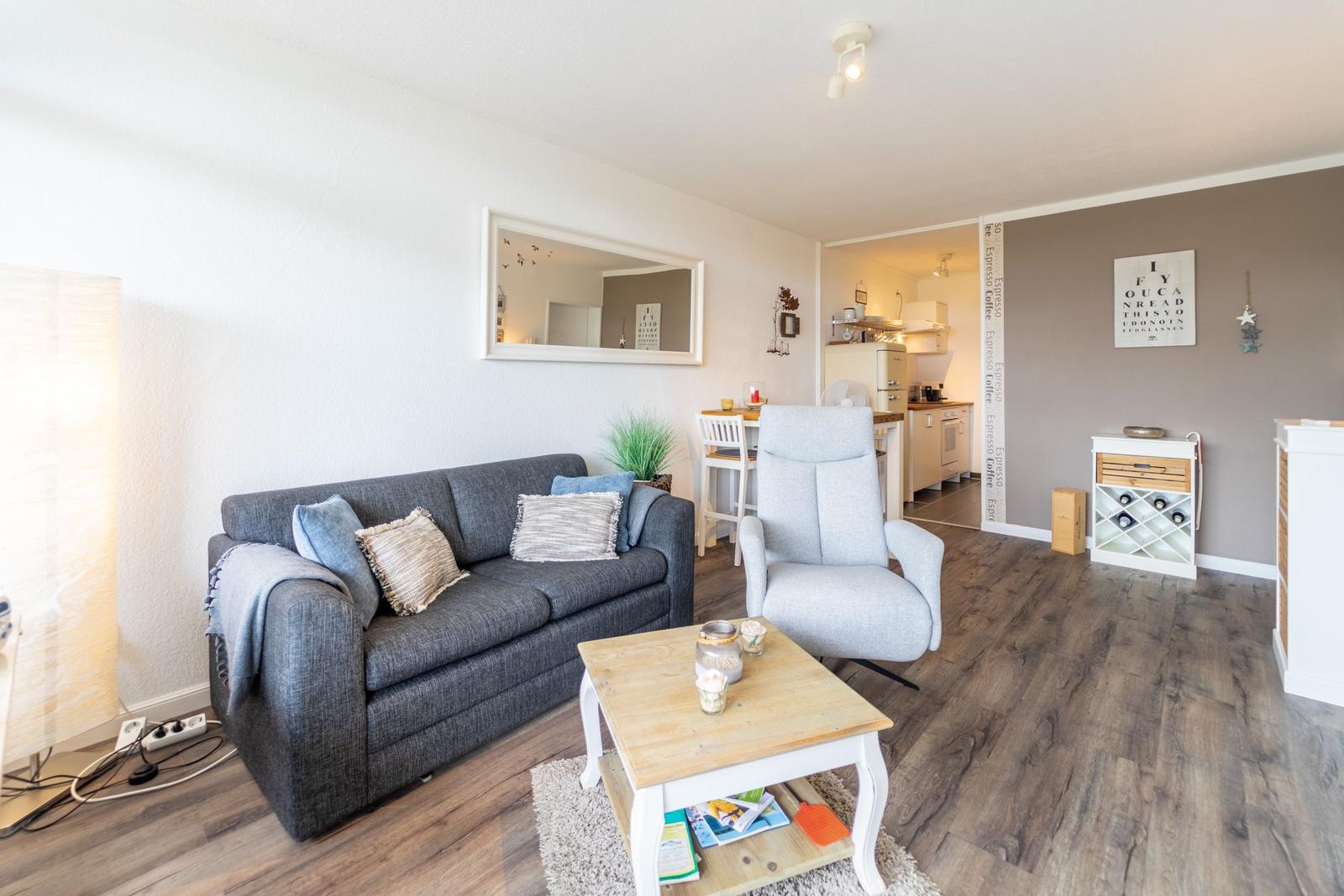 Wohnzimmer mit Sofa, Sessel und Couchtisch. Küche im Hintergrund.