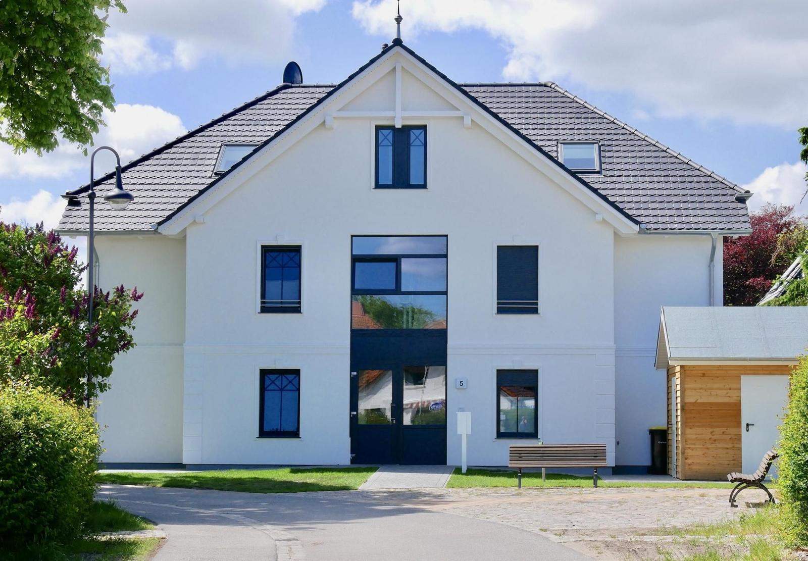 Weißes Haus mit dunklen Fenstern und Dach, Vorgarten mit Bank und Holzgarage.
