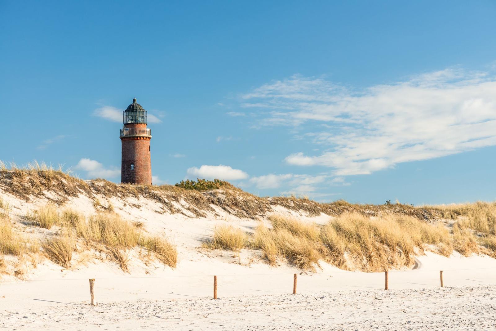 Roter Leuchtturm auf sandigem Strand mit Dünen und blauem Himmel.