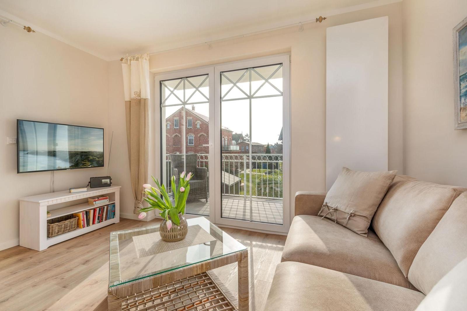 Wohnzimmer mit Sofa, Glas-Tisch und Balkontür. Fernseher und Bücherregal sichtbar.