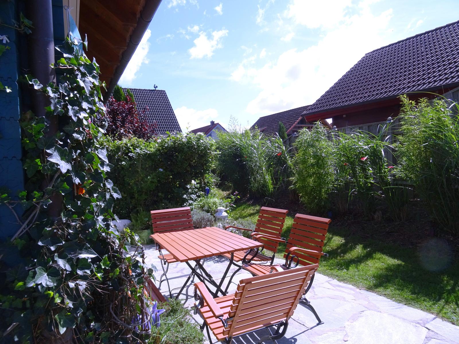 Terrasse mit Holztisch und Stühlen, umgeben von Grün und Häusern.