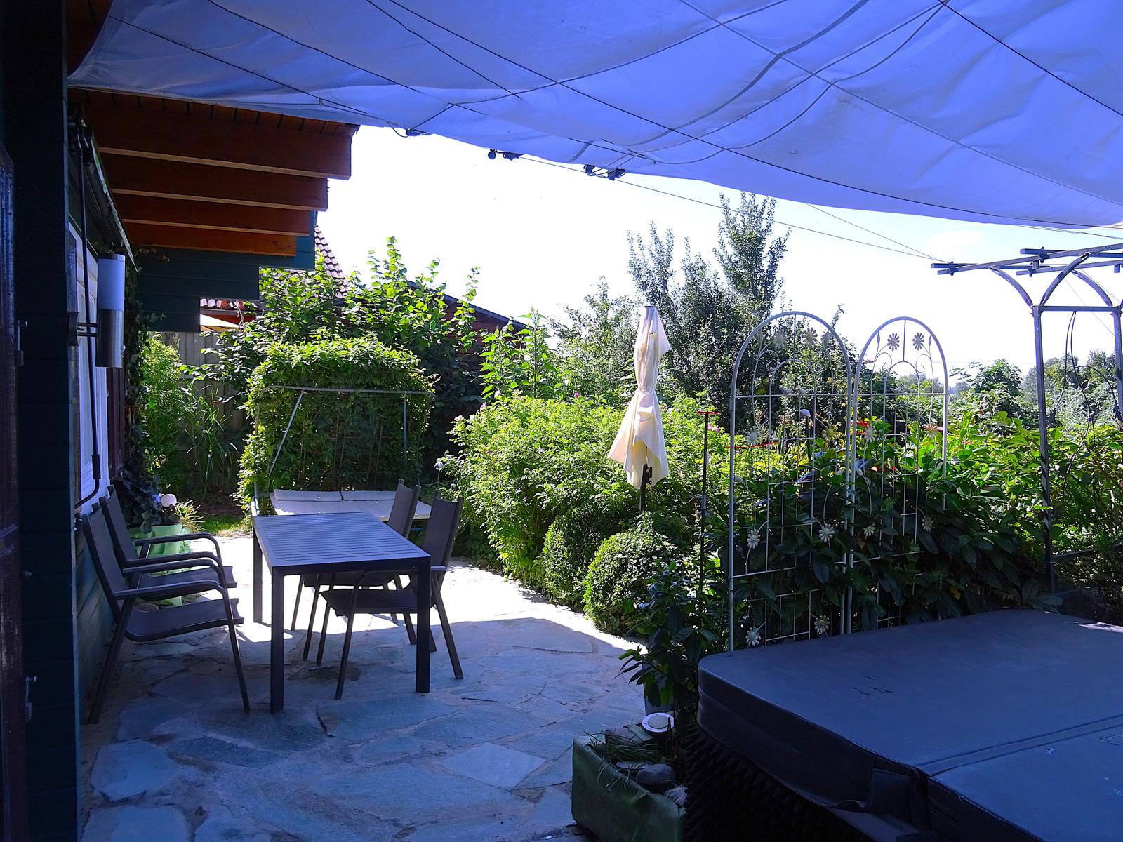 Terrasse mit Tisch, Stühlen und Sonnenschutz. Gartengrundstück mit Pflanzen und Gartenzaun.