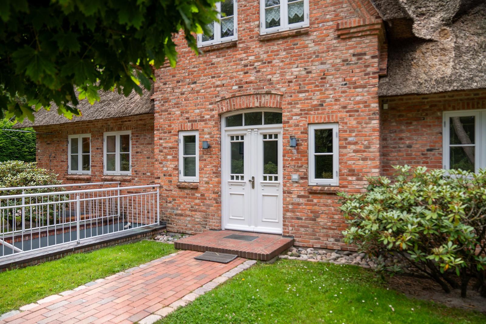Rotes Backsteinhaus mit Strohdach, weißer Eingangstür und gepflasterter Weg.