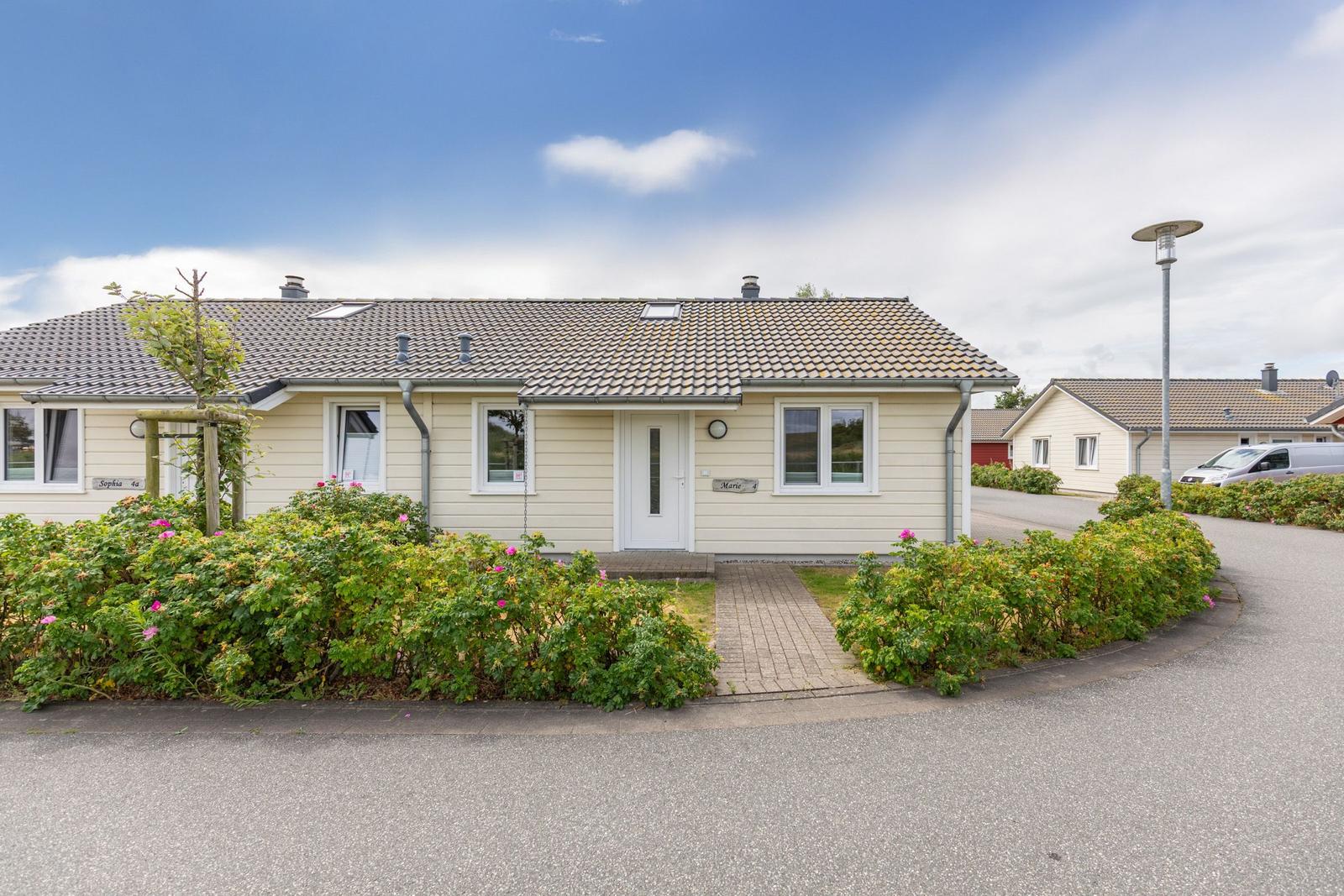 Ein hellfarbenes Haus mit Garten und gepflastertem Weg. Hintergrund: weitere Häuser und Straße.