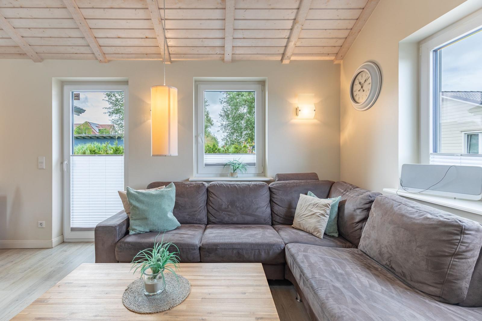 Wohnzimmer mit Ecksofa, Holztisch und Fenstern.