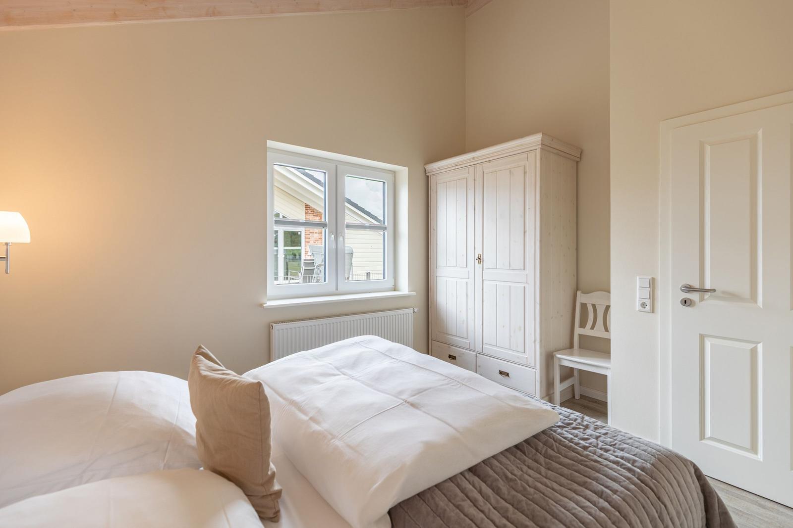 Schlafzimmer mit Bett, Kleiderschrank und Fenster. Tür und Stuhl sind sichtbar.