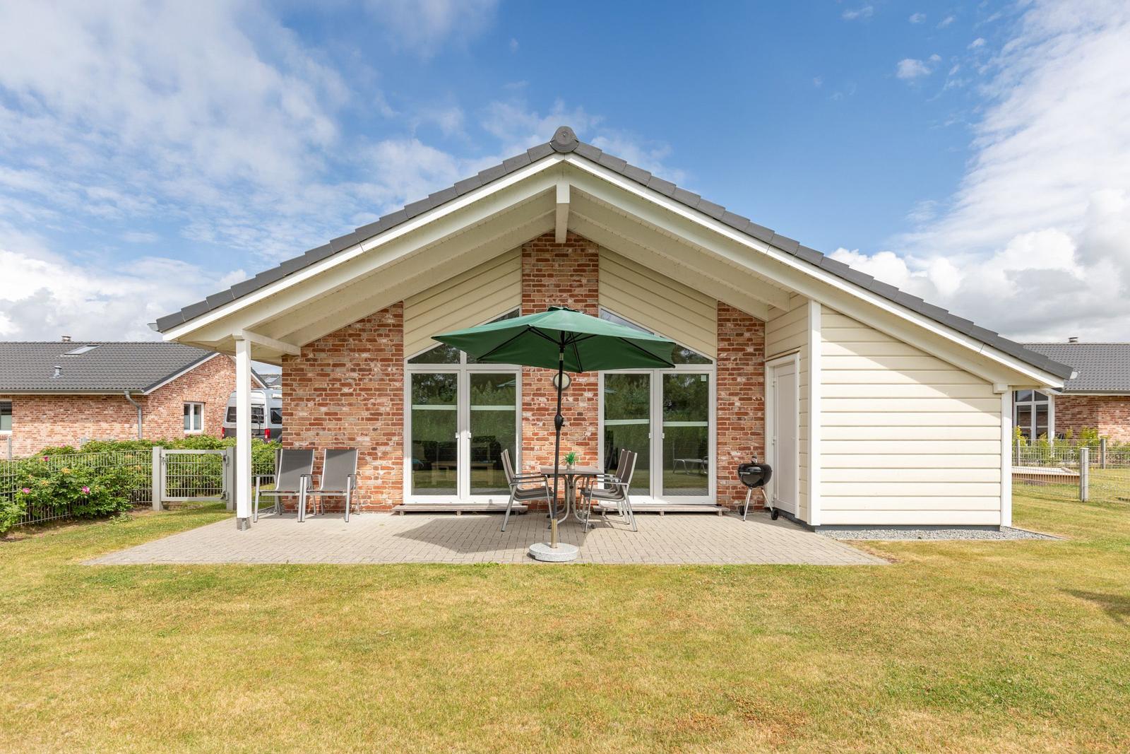 Haus mit Terrasse, grünem Sonnenschirm und Grill. Gartenbereich mit Sitzgelegenheiten.