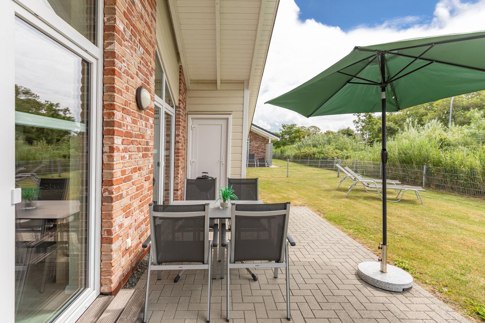 Terrasse mit Tisch, Stühlen und Sonnenschirm. Hinterhof mit Liegestühlen und Grünflächen.