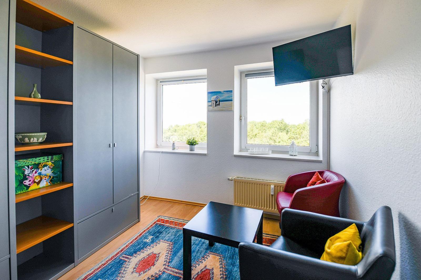 Zimmer mit Kleiderschrank, Sitzgelegenheit und TV. Fenster mit Blick auf Grün.