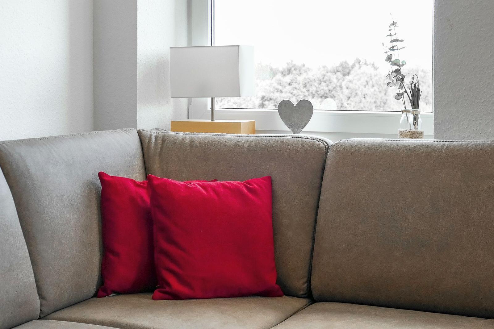Ein grauer Sofa mit zwei roten Kissen. Auf dem Fenster steht eine Lampe und ein Herz.