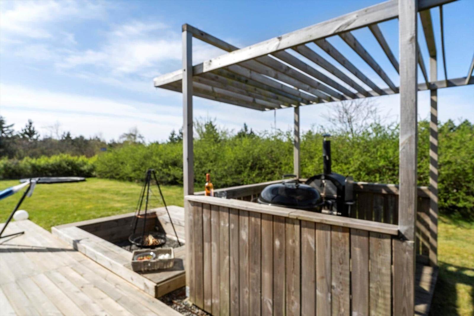 Holzterrasse mit Grill, Feuerkorb und Pavillon. Grünflächen und Bäume im Hintergrund.