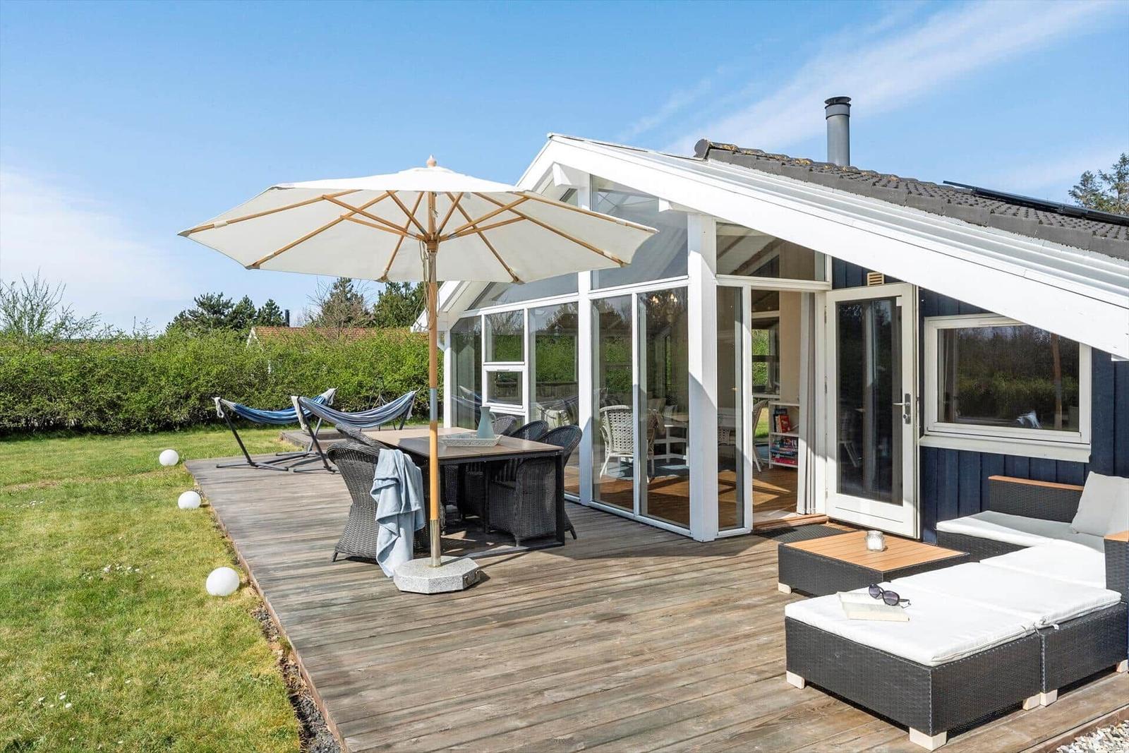 Terrasse mit Tisch, Stühlen und Sonnenschirm. Hintergrund: Haus mit Glasfront und Garten.