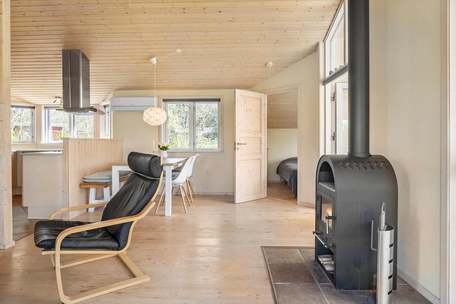 Holzvertäfelte Küche mit Esstisch, Sessel und Kaminofen. Schlafzimmer im Hintergrund.