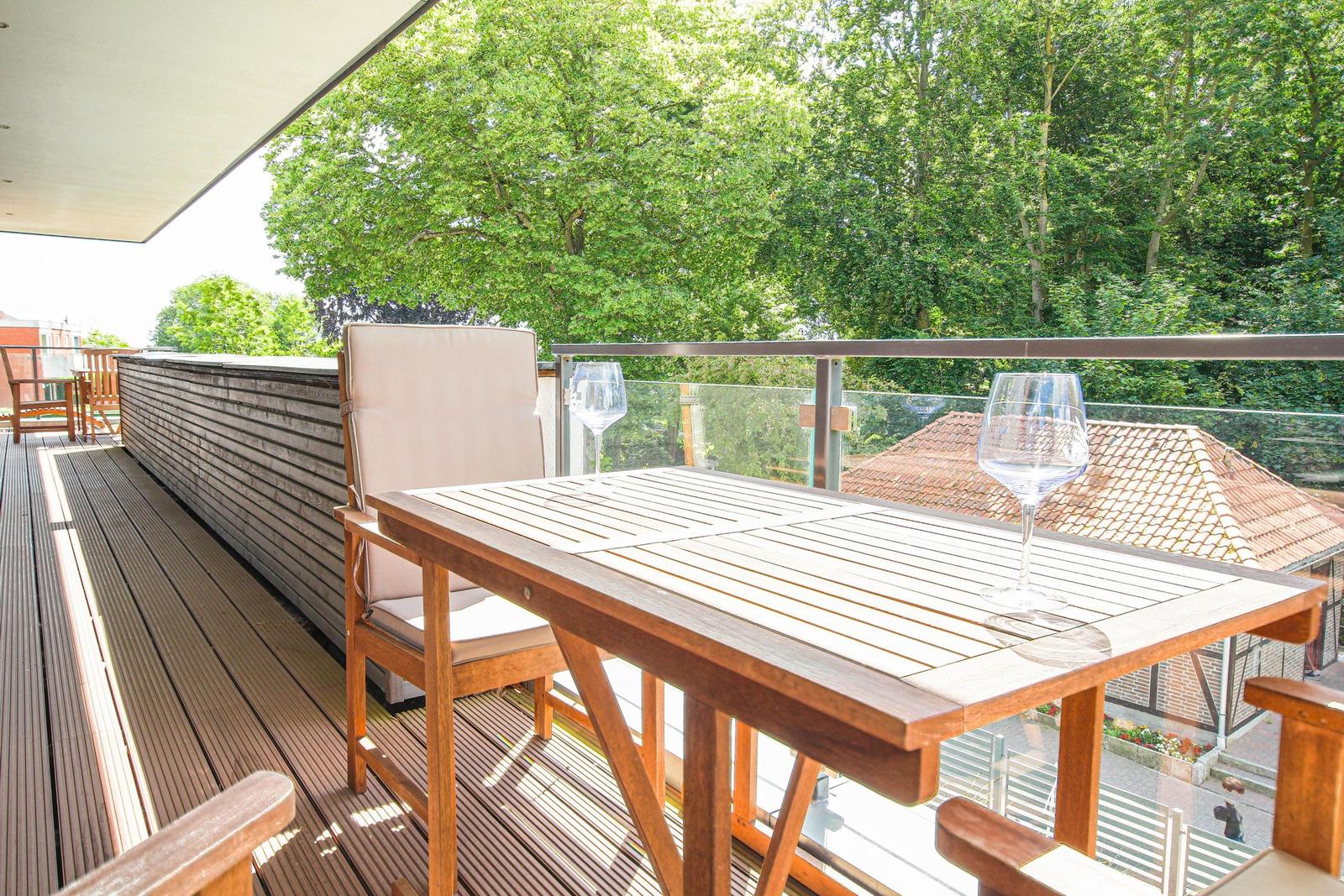 Terrasse mit Tisch, Stühlen und Weingläsern. Blick auf grüne Bäume und Dachziegel.