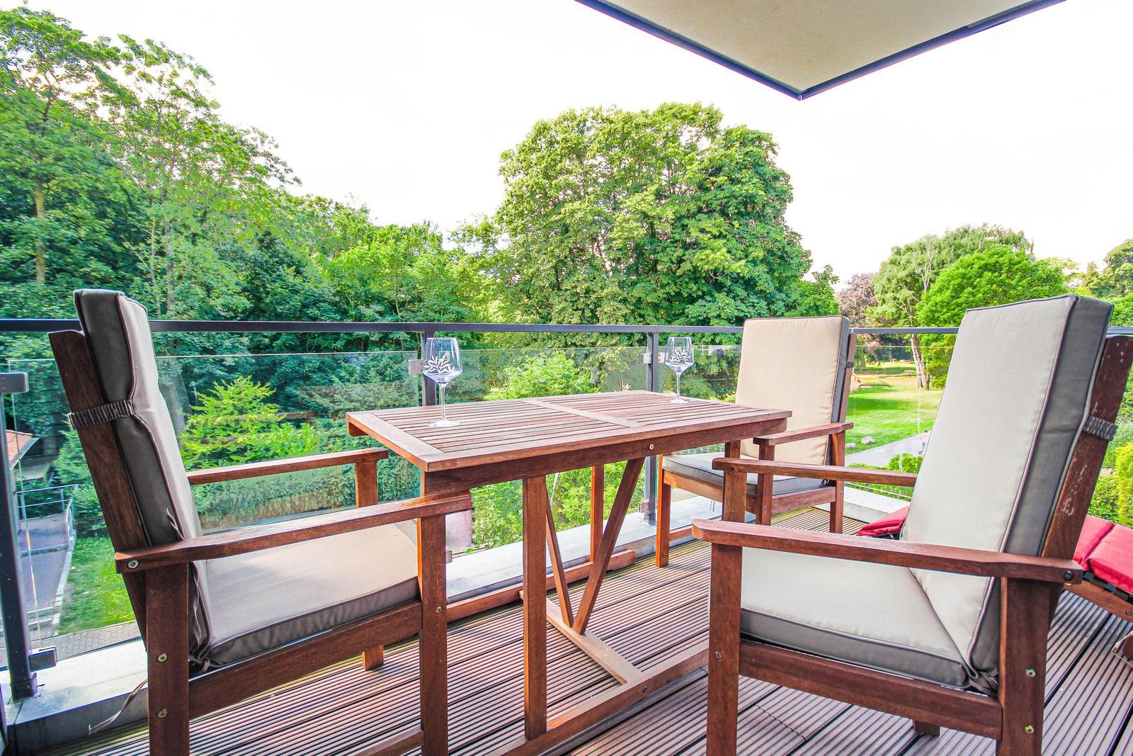 Terrasse mit Holztisch und Stühlen, Blick auf grüne Bäume und Rasen.