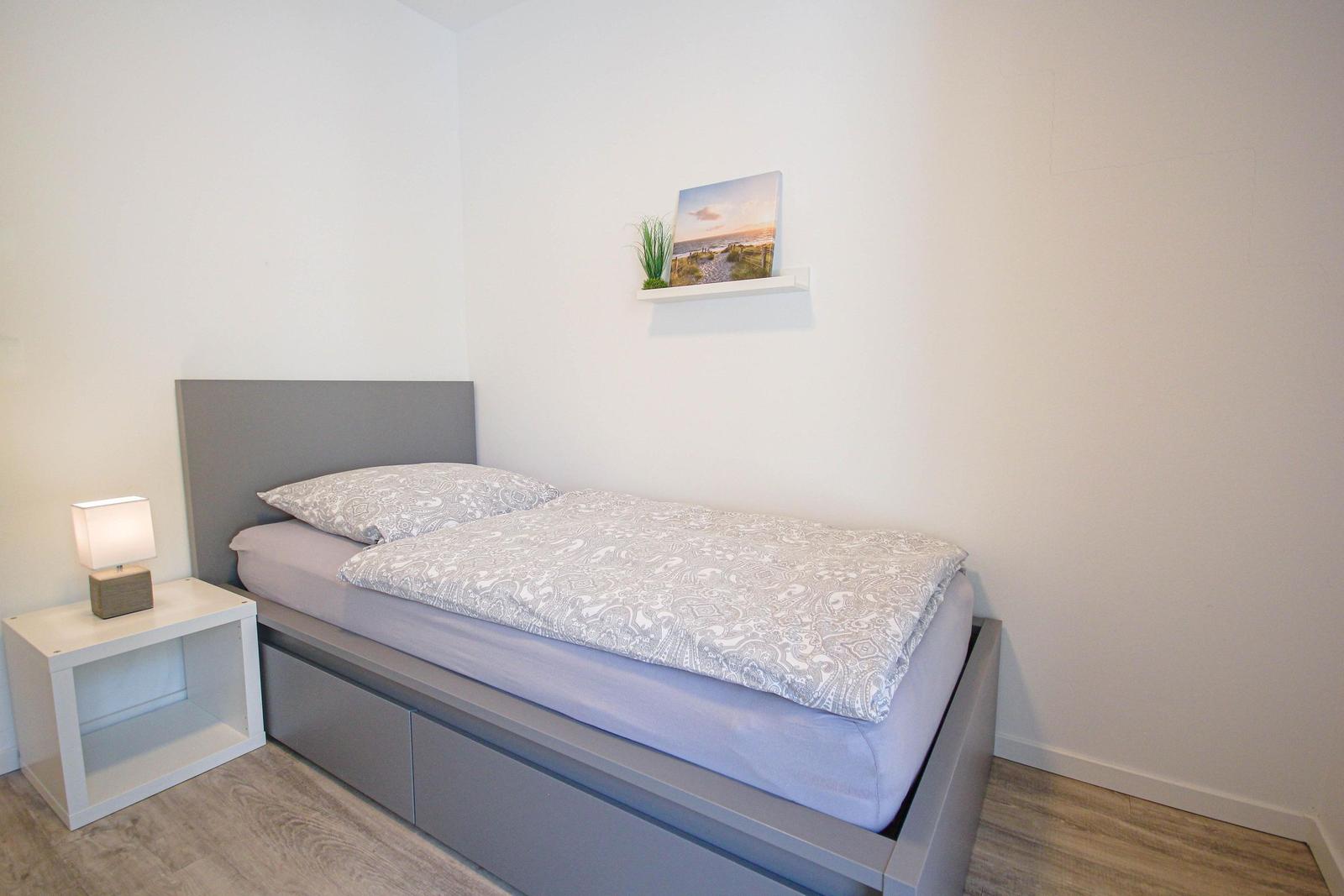 Schlafzimmer mit Bett, Nachttisch und Wandregal mit Bild und Pflanze.