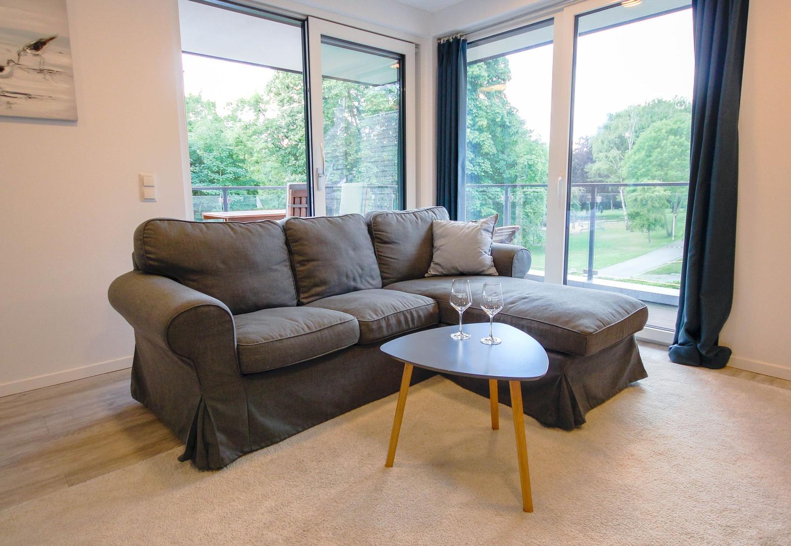 Ein Wohnzimmer mit grauem Sofa, Tisch und großen Fenstern zum Balkon.