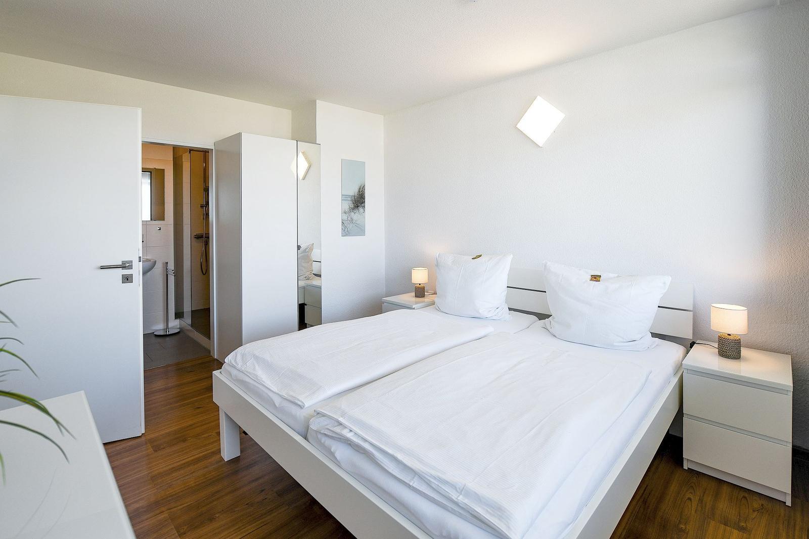 Schlafzimmer mit weißem Bett, Nachttischen und integriertem Bad.
