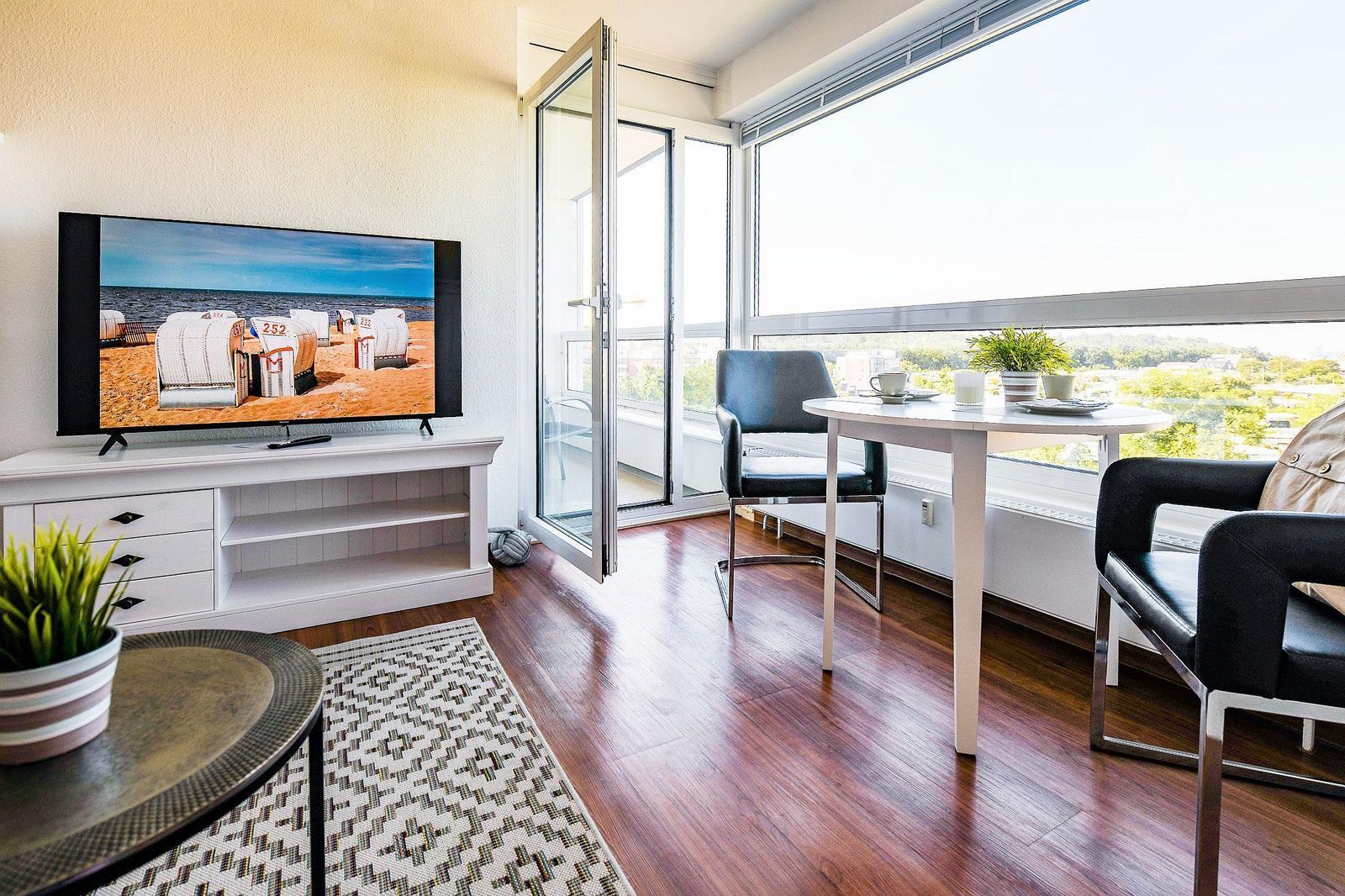 Wohnzimmer mit TV, Tisch und Stühlen. Balkon mit Aussicht. Holzboden und Teppich.