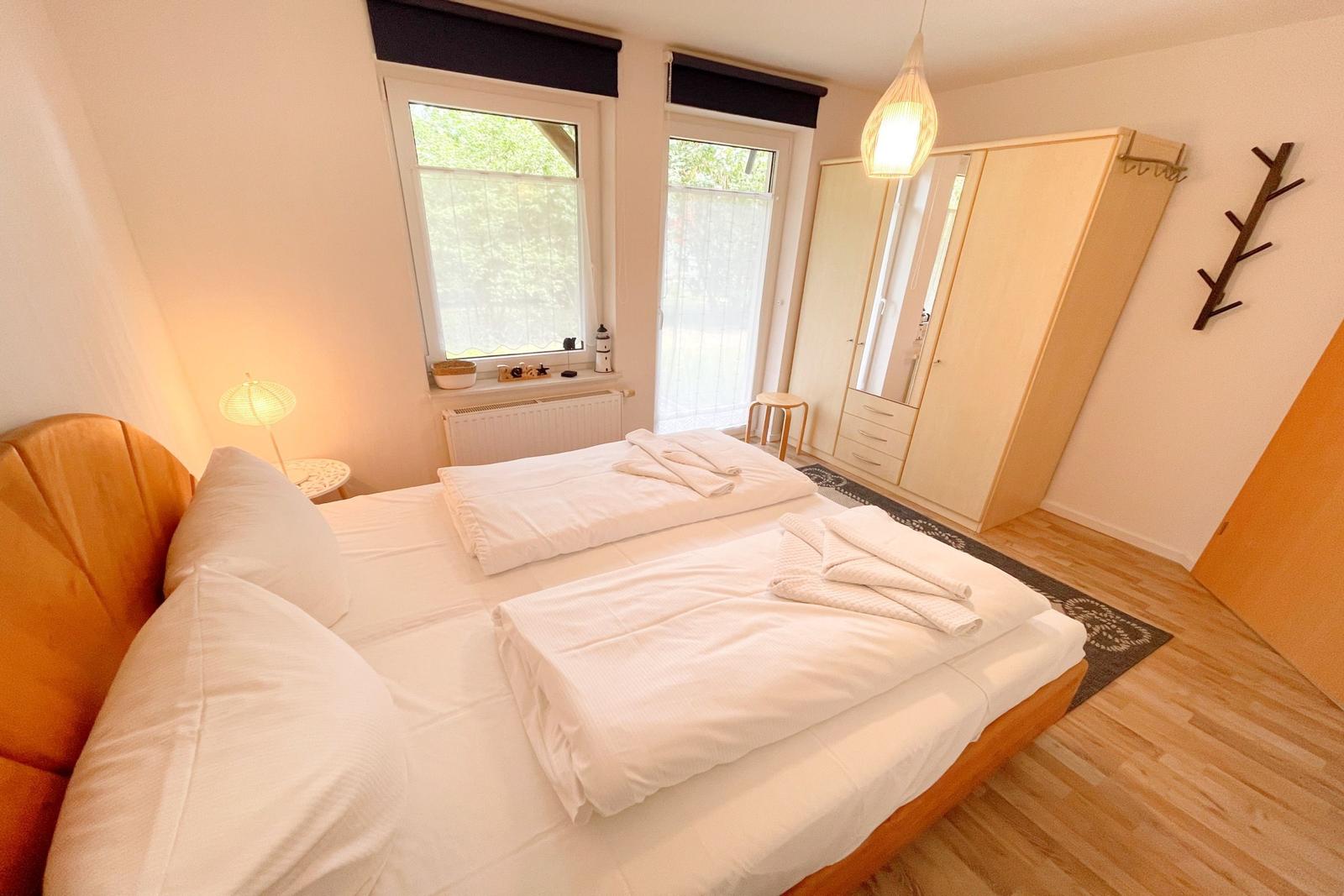 Doppelbett mit weißer Bettwäsche, Kleiderschrank und Fenster mit Blick auf Grün.