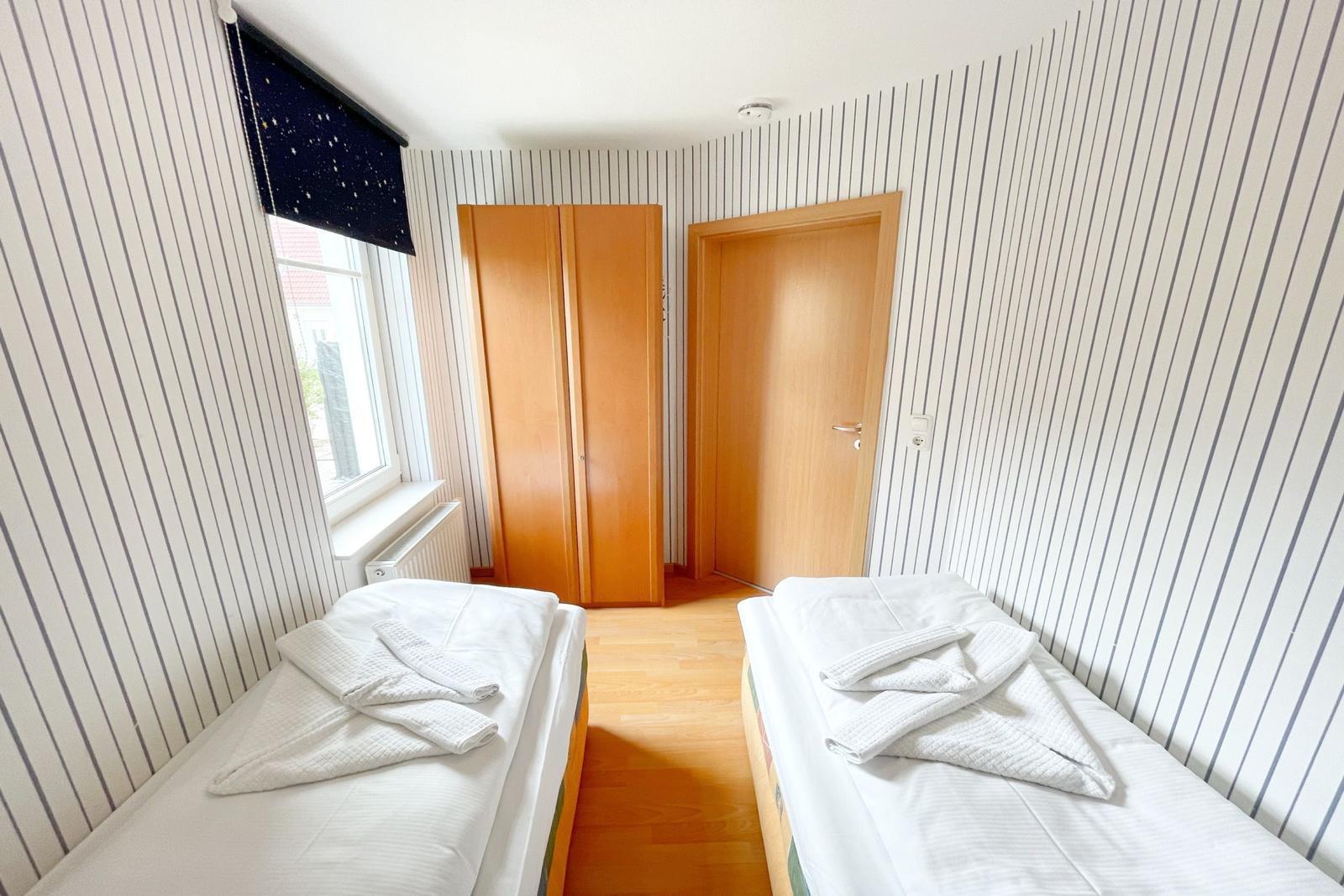 Zwei Betten mit weißen Laken und Handtüchern, Wand mit Streifen, Fenster und Tür.