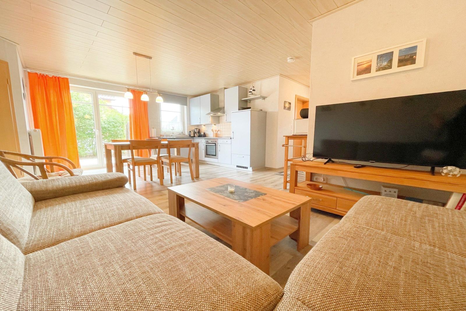 Wohnzimmer mit Sofa, Couchtisch und TV. Küche und Essbereich im Hintergrund.