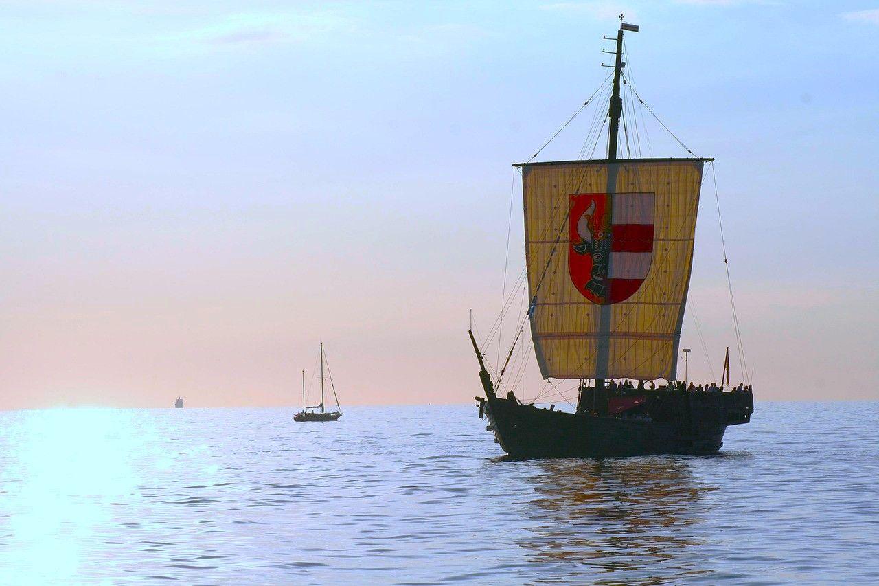 Historisches Segelschiff mit Wappen segelt auf ruhigem Meer. Im Hintergrund ist ein kleiner Segelboot sichtbar.