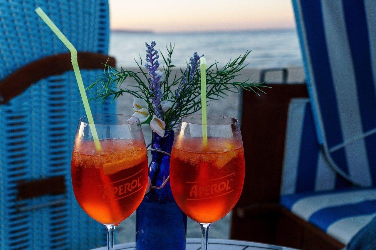 Zwei Aperol Spritz Getränke auf einem Tisch mit Meerblick und blauen Liegestühlen.