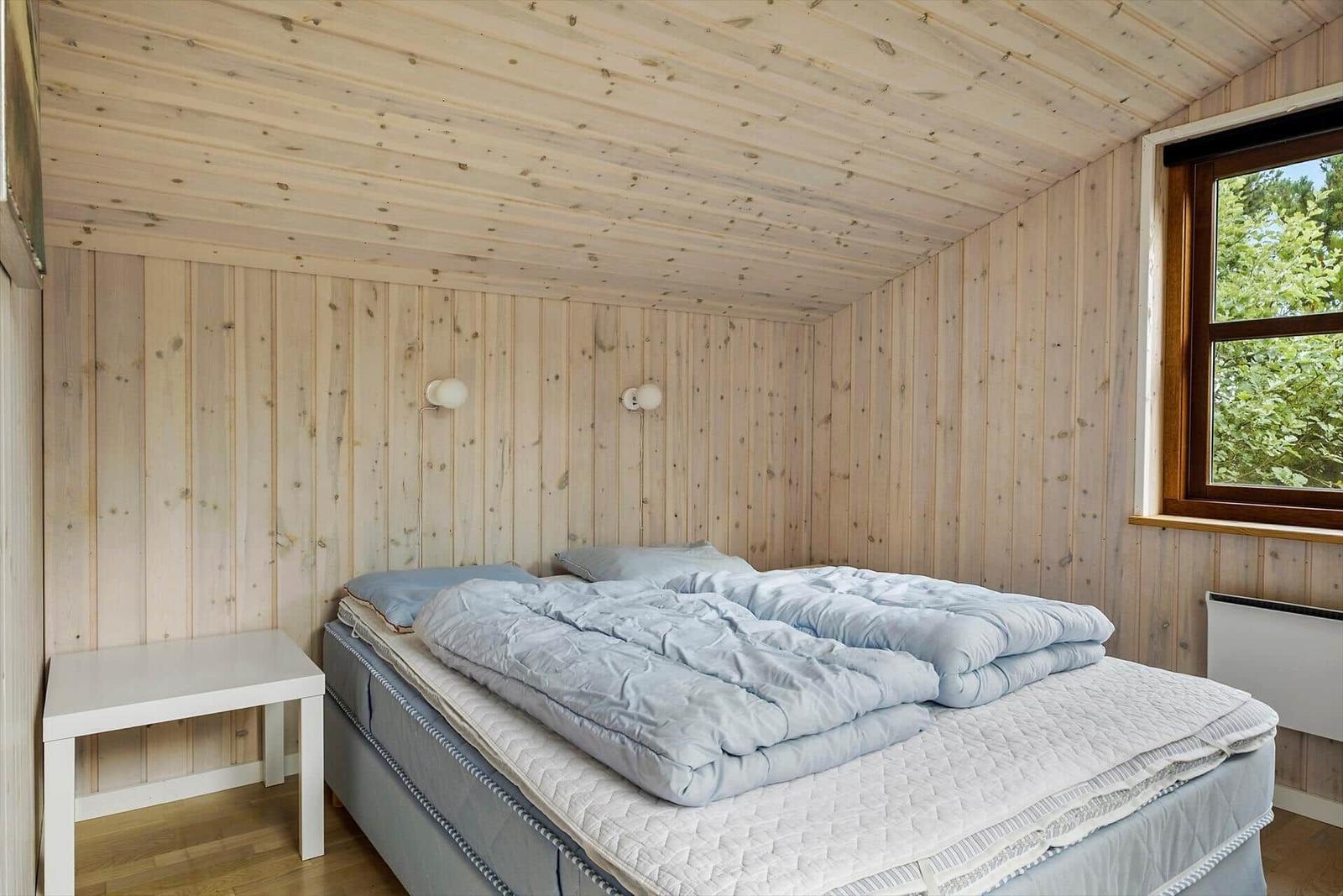 Schlafzimmer mit Holzwand, Bett und Fenster zur grünen Umgebung.