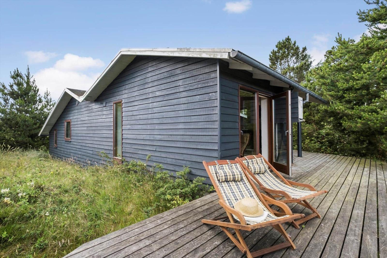 Holzterrasse mit zwei Liegestühlen vor einem grauen Holzhaus mit Glastür.