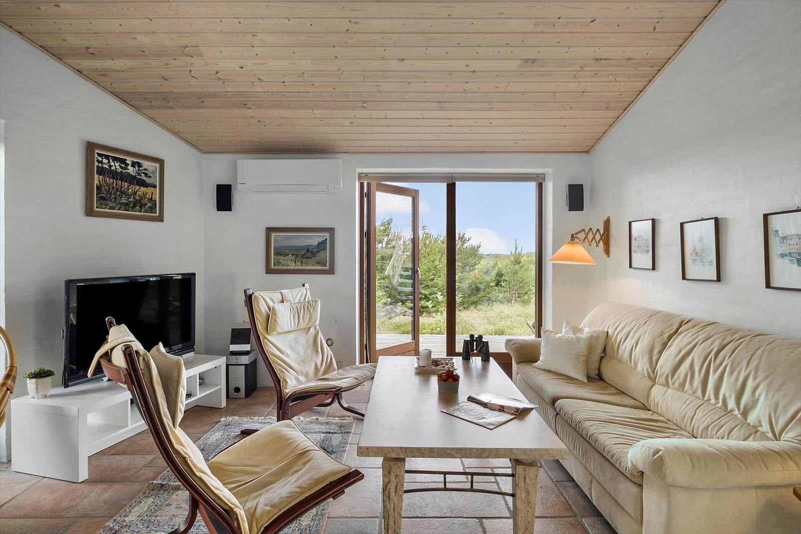 Wohnzimmer mit Sofa, Sessel und Fernseher. Durchgang zur Terrasse mit Blick auf Grünfläche.