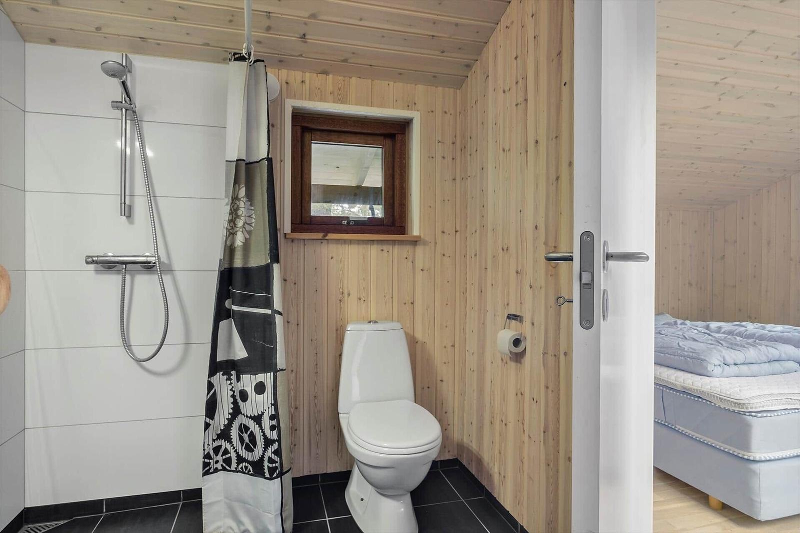 Badezimmer mit Dusche, Toilette und kleinem Fenster. Tür führt in Schlafzimmer mit Bett.