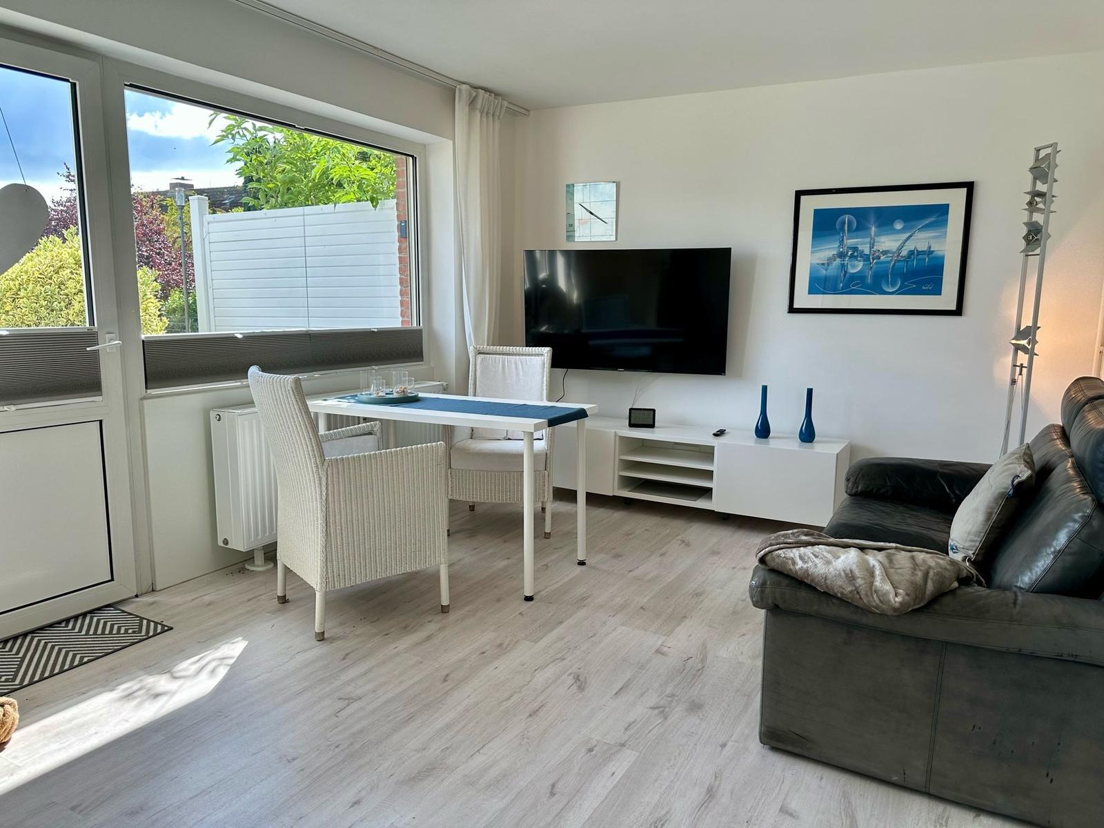 Wohn- und Essbereich mit Sofa, TV und Tisch. Große Fenster mit Blick auf Garten.