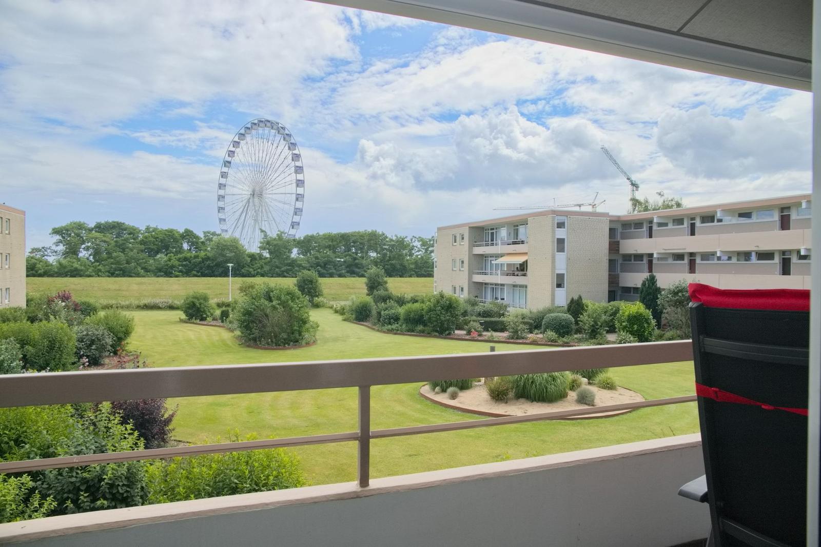 Balkon mit Sicht auf Grünfläche, Gebäude und Riesenrad unter blauem Himmel.