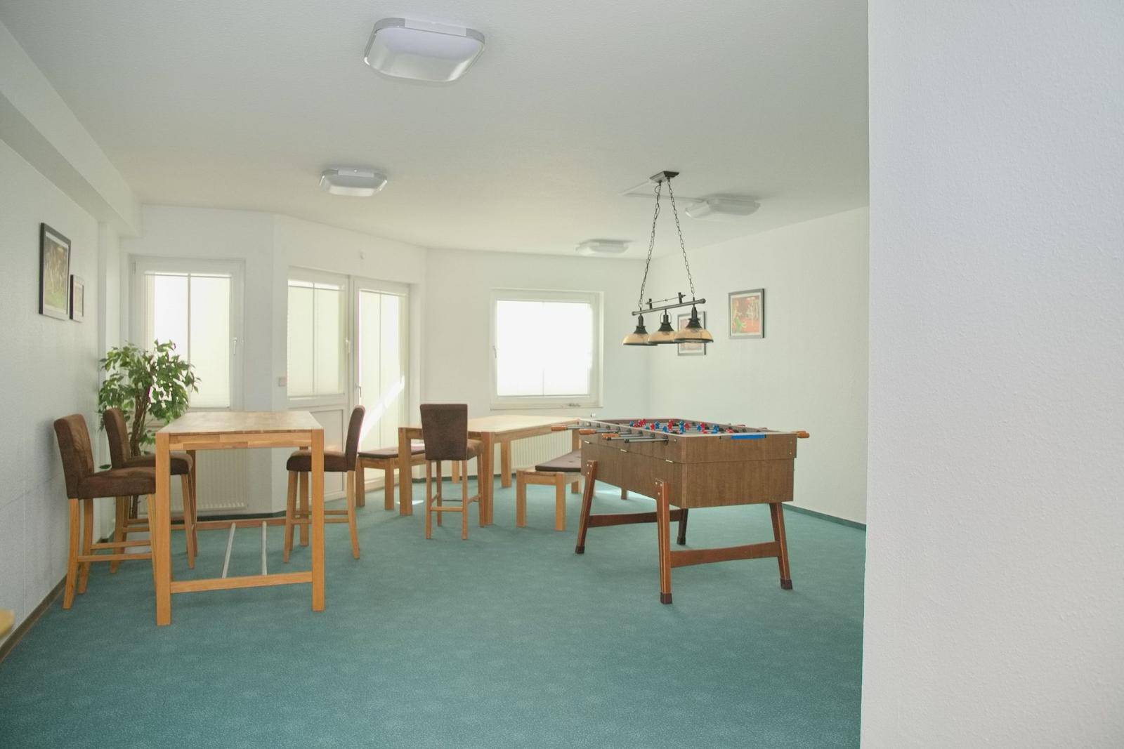 Großer Raum mit Tisch, Stühlen und Tischfußball. Blaues Teppichboden und Fenster.