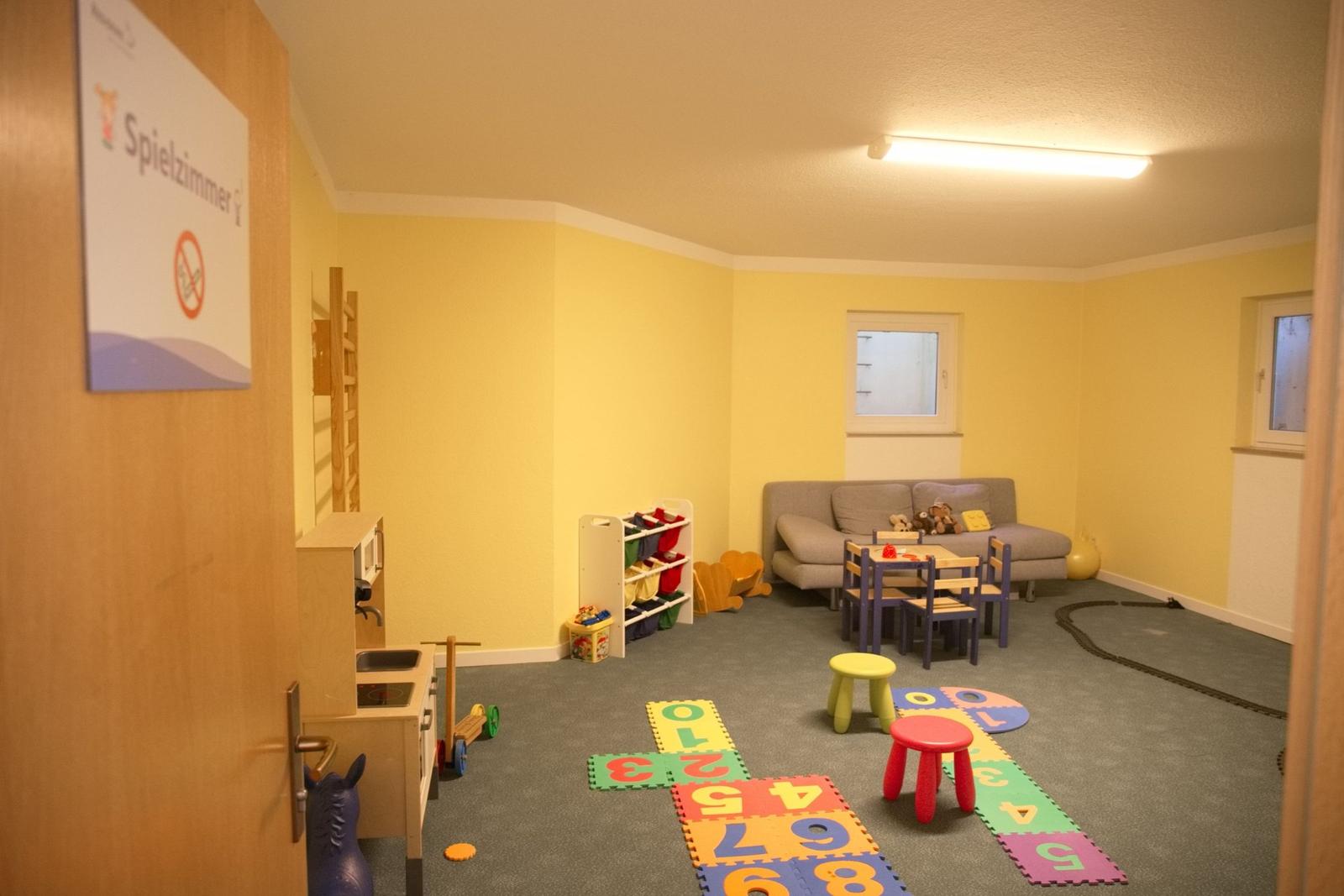Spielzimmer mit Kindersitzgruppe, Spielzeug und Zahlenmatte auf dem Boden.