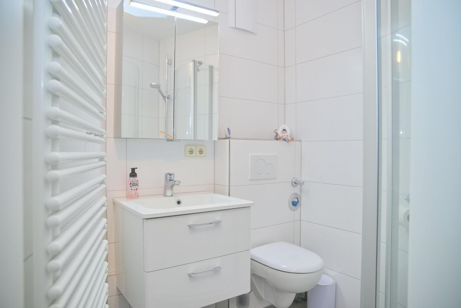 Badezimmer mit Waschbecken, Toilette und Dusche. Weiße Fliesen und Spiegelschrank.