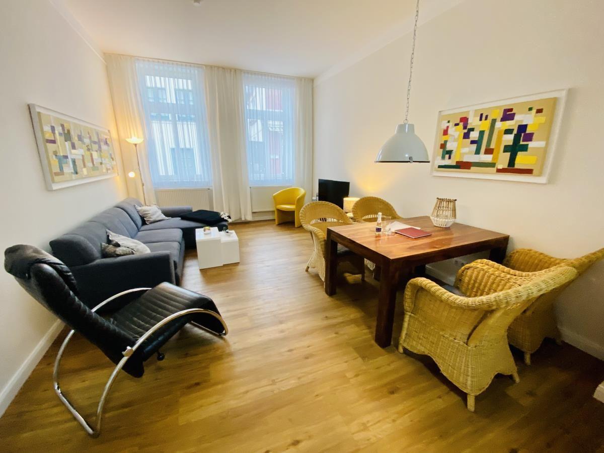 Wohn- und Essbereich mit Sofa, Esstisch und Stühlen. Holzboden und Fenster mit Vorhängen.