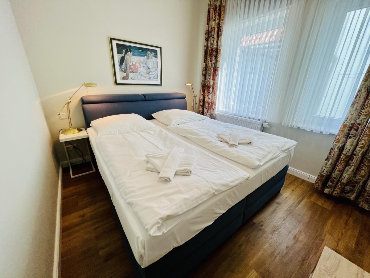 Doppelbett mit weißen Bettwäsche, Holzboden und Fenster mit Vorhängen.