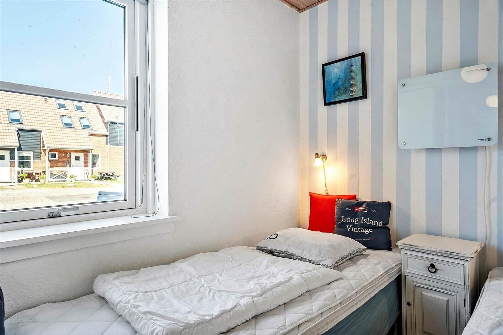 Ein Schlafzimmer mit Bett, Nachttisch und Fenster mit Blick auf Häuser.