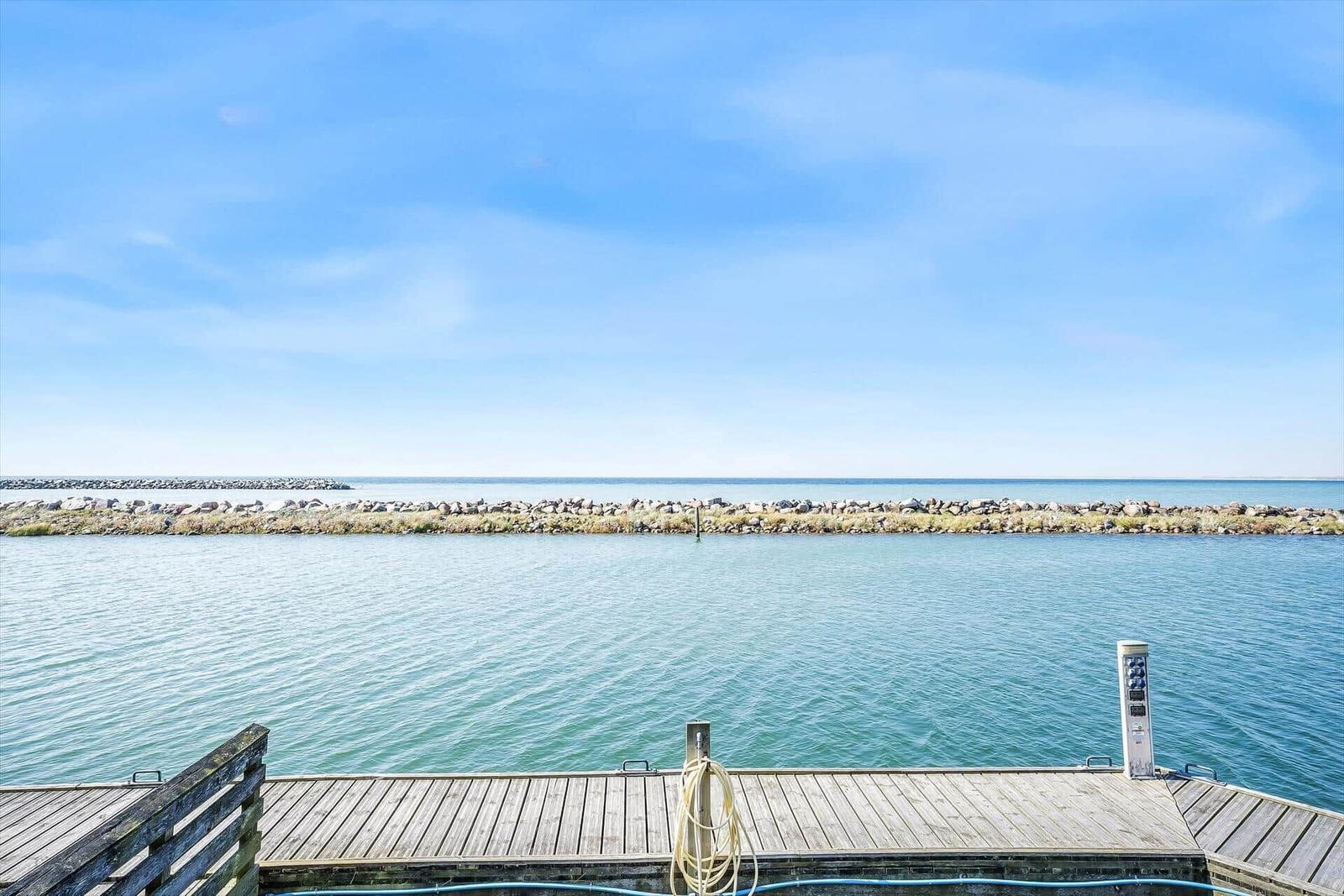 Holzsteg mit Blick auf ruhiges Wasser und Steinufer unter blauem Himmel.
