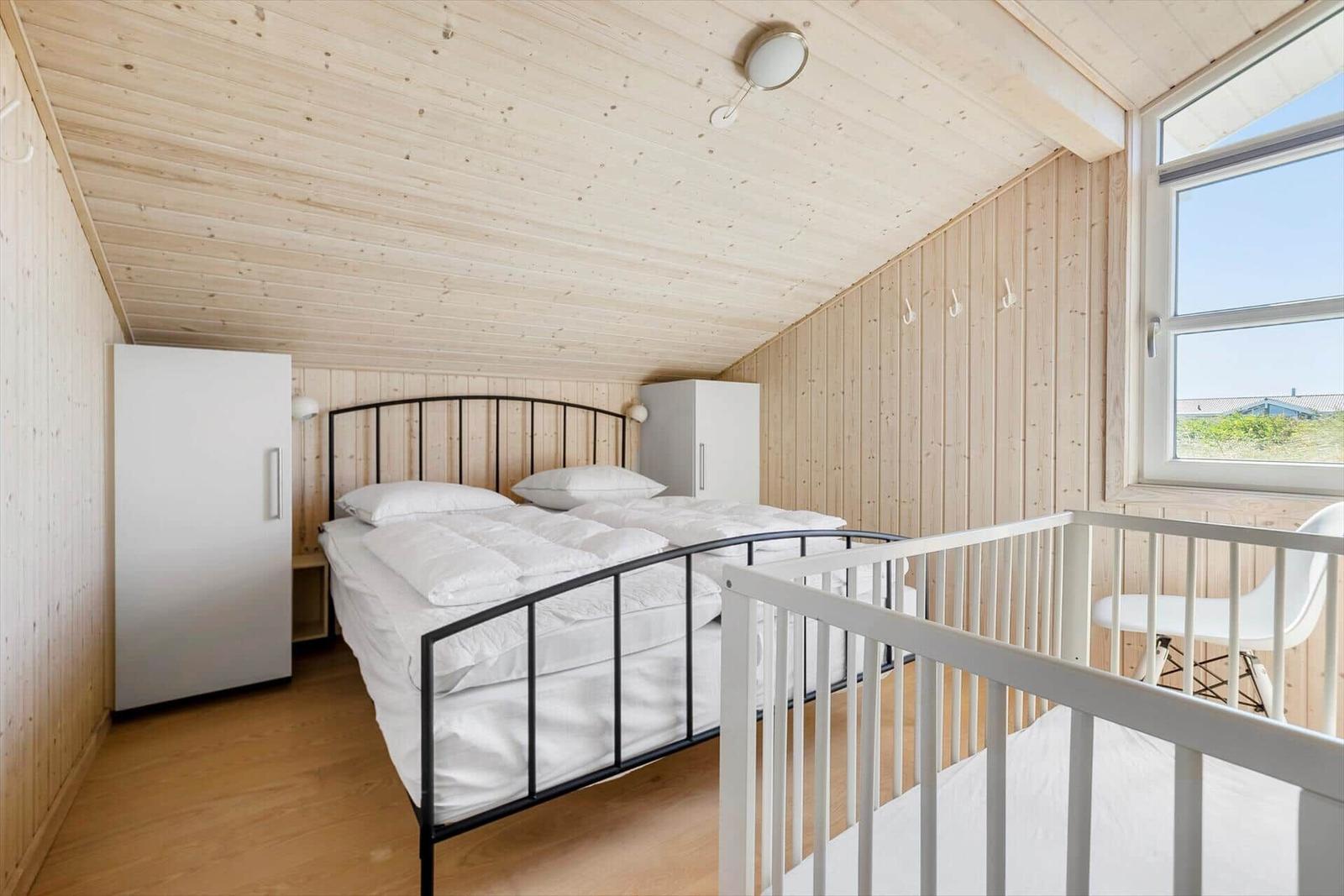 Schlafzimmer mit Doppelbett, weißen Schränken und Holzwänden. Fenster mit Sicht auf Außengelände.