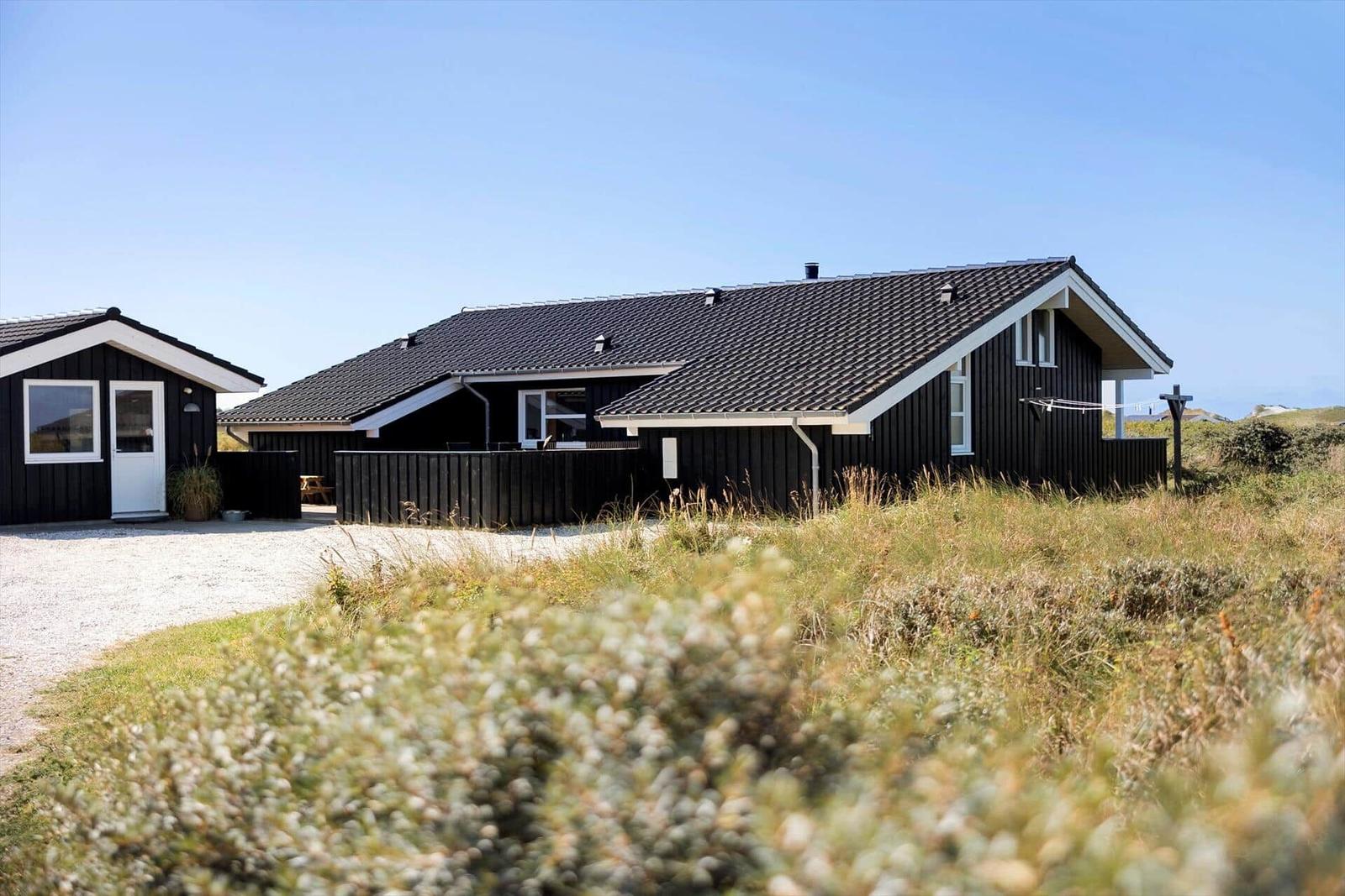 Schwarzes Holzhaus mit weißem Fensterrahmen und Dach. Umgeben von Gras und einem Kiesweg.