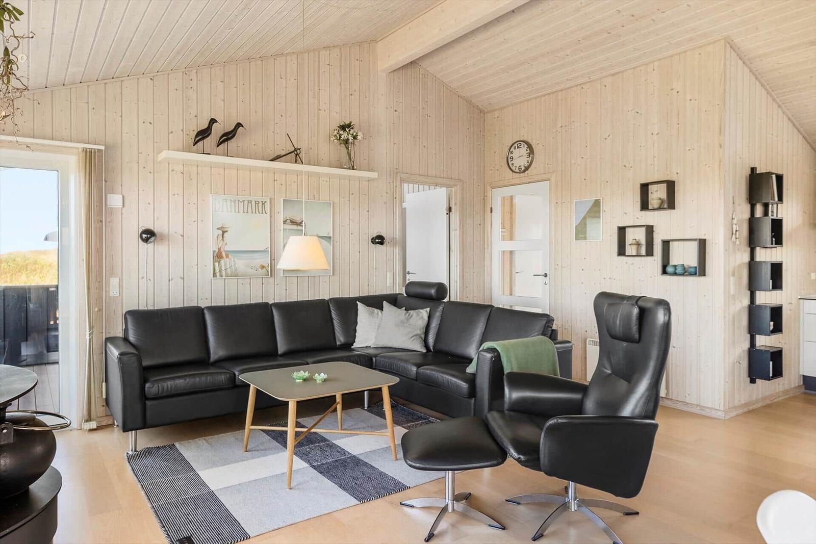 Wohnzimmer mit schwarzer Ledercouch, Sessel und Holzmöbeln an Holzwand.
