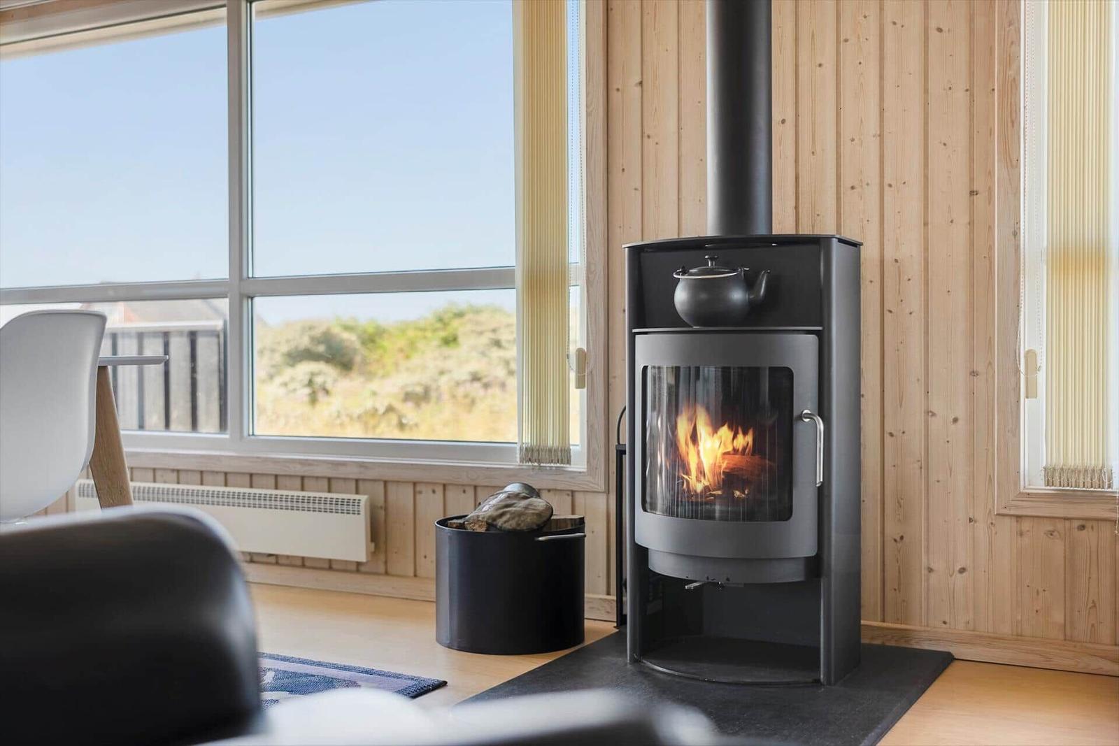 Kaminofen mit brennendem Holz und Teekanne, Holzwand, Fenster mit Blick nach draußen.