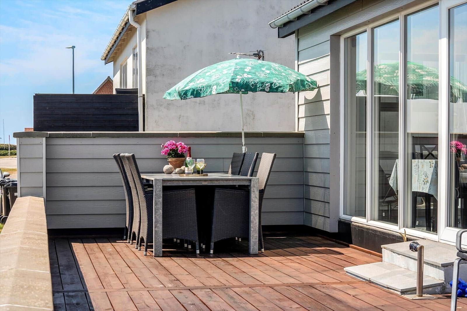 Terrasse mit Tisch, Stühlen und grünem Sonnenschirm. Holzdecke und Glasfassade sichtbar.